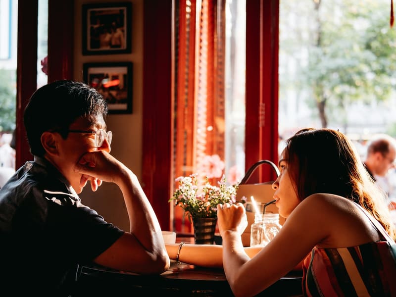 Two people talking across a café table by a sunlit window.