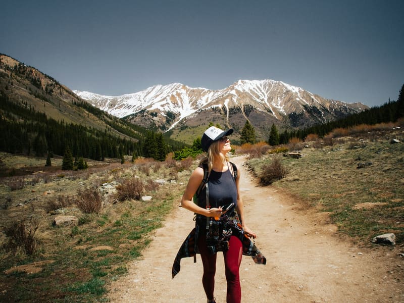 A hiker on a dirt trail with snow-capped mountains behind her.